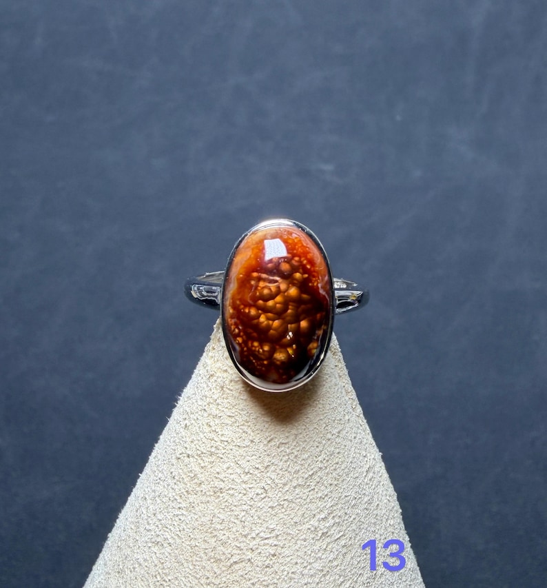 Op de afbeelding: Een zilveren ring met een ovaalvormige cabochonsteen met een donkeroranje en bruin gevlekt patroon. De steen heeft een glanzend, getextureerd oppervlak met lichtere gouden vlekjes. Het getal 13 staat in het blauw rechtsonder gedrukt.