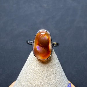 Op de afbeelding: Een zilverkleurige ring met een gladde, gepolijste, ovaalvormige cabochonsteen in een diepe roodoranje tint met iriserende lichtflitsen. De steen is gezet in een eenvoudige metalen zetting en het cijfer 4 is zichtbaar in de benedenhoek.