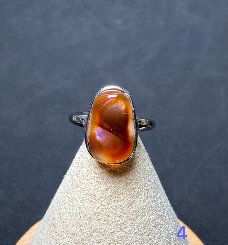 Op de afbeelding: Een zilverkleurige ring met een gladde, gepolijste, ovaalvormige cabochonsteen in een diepe roodoranje tint met iriserende lichtflitsen. De steen is gezet in een eenvoudige metalen zetting en het cijfer 4 is zichtbaar in de benedenhoek.