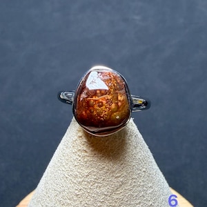 Op de afbeelding: Een ring met een gepolijste, onregelmatige, dieproodbruine steen met gouden, bubbelachtige insluitingen. De steen is gezet in een gladde, zilverkleurige metalen zetting en ringband, wat zorgt voor een reflecterend oppervlak en een organische vorm.