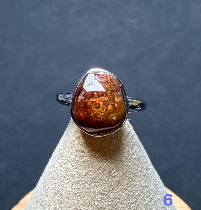 Op de afbeelding: Een ring met een gepolijste, onregelmatige, dieproodbruine steen met gouden, bubbelachtige insluitingen. De steen is gezet in een gladde, zilverkleurige metalen zetting en ringband, wat zorgt voor een reflecterend oppervlak en een organische vorm.