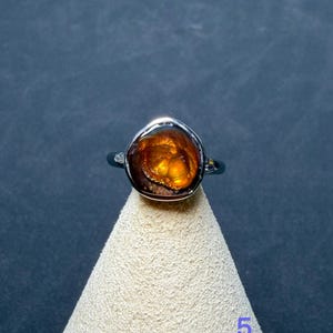 Op de afbeelding: Een zilveren ring met een gepolijste, onregelmatige vuuragaat edelsteen in cabochon-slijpvorm met diepe oranje en bruine tinten. De steen vertoont een gloeiende, iriserende interne structuur in een gladde metalen zetting.