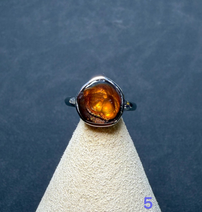 Op de afbeelding: Een zilveren ring met een gepolijste, onregelmatige vuuragaat edelsteen in cabochon-slijpvorm met diepe oranje en bruine tinten. De steen vertoont een gloeiende, iriserende interne structuur in een gladde metalen zetting.