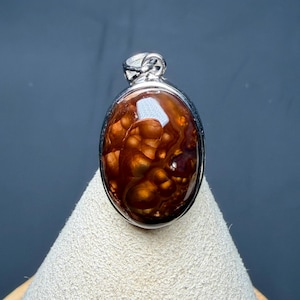 May include: An oval-shaped pendant featuring a polished fire agate gemstone with deep reddish-brown and orange iridescent patterns, encased in a smooth silver-toned metal setting with a small bail at the top for hanging.