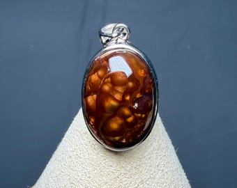 Op de afbeelding: Een ovaalvormige hanger met een gepolijste vuuragaat edelsteen met dieproodbruine en oranje iriserende patronen, gevat in een gladde zilverkleurige metalen zetting met een klein oogje aan de bovenzijde om op te hangen.