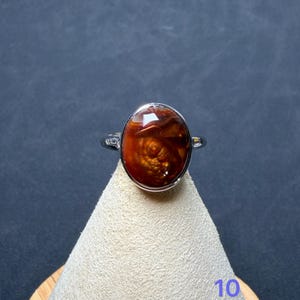 Op de afbeelding: Een zilverkleurige ring met een grote, ovale, dieproodbruine steen met een glad, gepolijst oppervlak en interne wervelpatronen. De ring wordt gepresenteerd op een lichtgekleurde, conische standaard met het cijfer 10 zichtbaar.