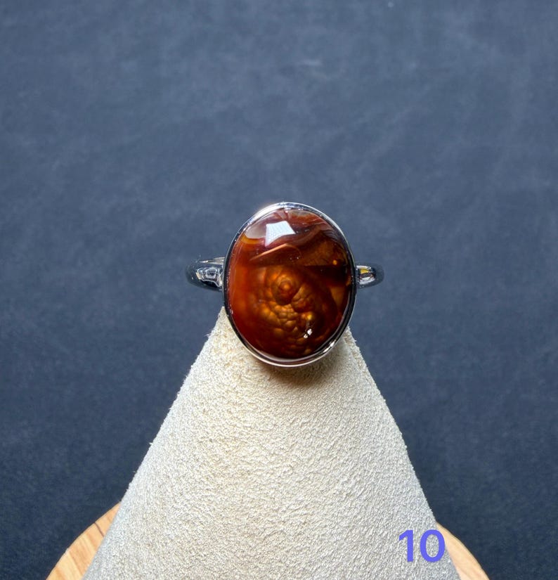 Op de afbeelding: Een zilverkleurige ring met een grote, ovale, dieproodbruine steen met een glad, gepolijst oppervlak en interne wervelpatronen. De ring wordt gepresenteerd op een lichtgekleurde, conische standaard met het cijfer 10 zichtbaar.