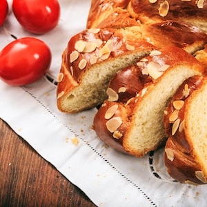 May include: A braided loaf of sweet bread topped with sliced almonds, cut into slices, on a white tablecloth with red Easter eggs.