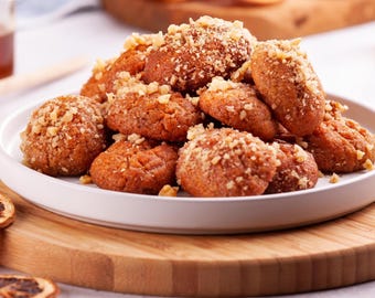 May include: A plate of traditional Greek Melomakarona cookies, a festive treat. These oval-shaped cookies are golden brown, topped with chopped nuts, and served on a white plate atop a wooden board. Honey and dried orange slices are also visible.
