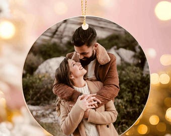 May include: A round Christmas ornament with a photo of a couple embracing. The ornament is suspended by a gold string and is surrounded by a blurred background of a Christmas tree with white frosted branches and warm lights.