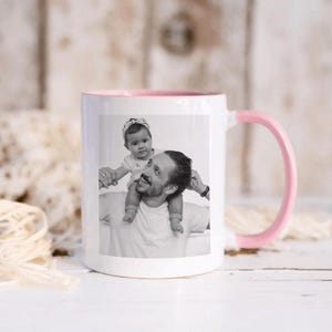 May include: White ceramic mug with a pink interior and handle. The mug features a black and white photograph of a person carrying a baby on their shoulders. A perfect gift for any occasion.