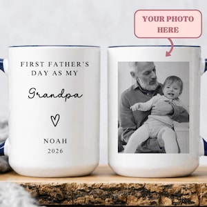 May include: White ceramic mug with a navy blue handle and rim. The mug features the text "FIRST FATHER'S DAY AS MY Grandpa" with a heart symbol and the name "NOAH" and the year "2026". The other side has a black and white photo.