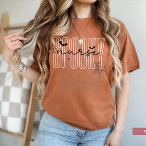 Puede incluir: Camiseta color &oacute;xido con la frase "SPOOKY nurse" en blanco, con murci&eacute;lagos y telara&ntilde;as. Esta camiseta de estilo casual es perfecta para Halloween o como regalo para una enfermera. Se combina con vaqueros azules.