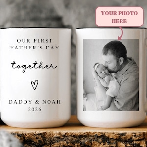 May include: White ceramic mug with a black handle and rim. The mug features the text "OUR FIRST FATHER'S DAY together" with a heart symbol and "DADDY & NOAH 2026". The other side displays a black and white photo of a father and baby. The text "YOUR PHOTO HERE" is above the photo.