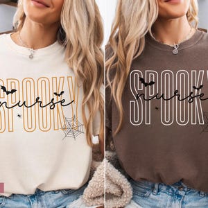 Puede incluir: Dos camisetas, una marfil y otra marr&oacute;n, con el texto "Spooky Nurse" en un dise&ntilde;o de Halloween. El dise&ntilde;o incluye murci&eacute;lagos, una ara&ntilde;a y una telara&ntilde;a. La camiseta marfil tiene texto naranja, la camiseta marr&oacute;n tiene texto blanco.