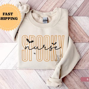 Puede incluir: Sudadera color arena con las palabras "Spooky Nurse" en letras naranjas y negras. El dise&ntilde;o incluye murci&eacute;lagos, una ara&ntilde;a y una telara&ntilde;a, ideal para Halloween. La sudadera est&aacute; doblada cuidadosamente.
