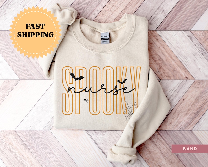 Puede incluir: Sudadera color arena con las palabras "Spooky Nurse" en letras naranjas y negras. El dise&ntilde;o incluye murci&eacute;lagos, una ara&ntilde;a y una telara&ntilde;a, ideal para Halloween. La sudadera est&aacute; doblada cuidadosamente.