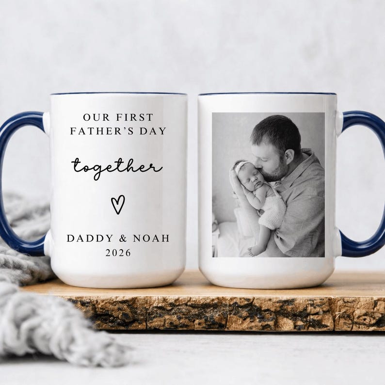 May include: White ceramic mug with a navy blue handle and rim. The mug features the text "OUR FIRST FATHER'S DAY together" with a heart symbol and "DADDY & NOAH 2026". The other side displays a black and white photo of a father holding a baby.