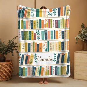 May include: A white blanket featuring a colorful bookshelf design with rows of books in various colors, including blue, orange, and green. The name "Camila" is printed in cursive. Floral and leaf accents are also present.