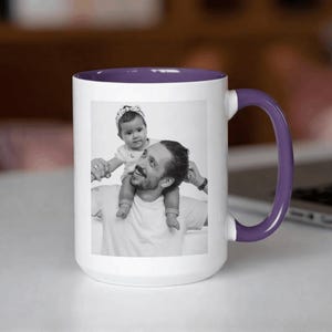 May include: White ceramic mug with a purple interior and handle. The mug features a black and white photograph of a person with a baby on their shoulders. The mug is sitting on a white surface.