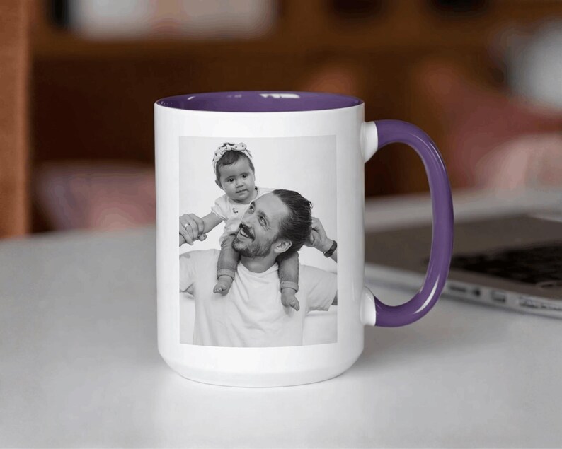 May include: White ceramic mug with a purple interior and handle. The mug features a black and white photograph of a person with a baby on their shoulders. The mug is sitting on a white surface.