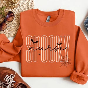 Puede incluir: Sudadera naranja con la frase "SPOOKY nurse" en blanco y negro. El dise&ntilde;o incluye murci&eacute;lagos, una ara&ntilde;a y una telara&ntilde;a, perfecto para Halloween. La sudadera se combina con vaqueros y zapatos marrones.