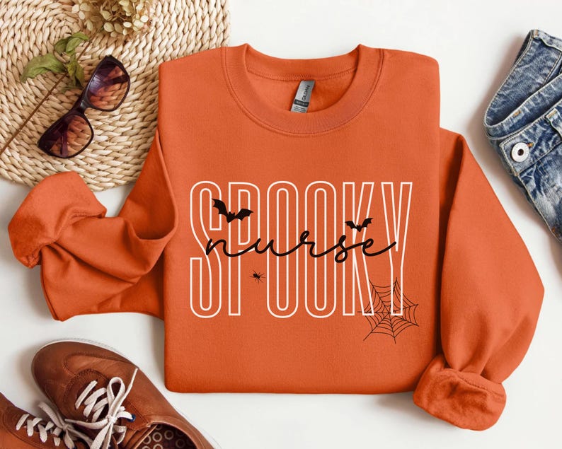 Puede incluir: Sudadera naranja con la frase "SPOOKY nurse" en blanco y negro. El dise&ntilde;o incluye murci&eacute;lagos, una ara&ntilde;a y una telara&ntilde;a, perfecto para Halloween. La sudadera se combina con vaqueros y zapatos marrones.