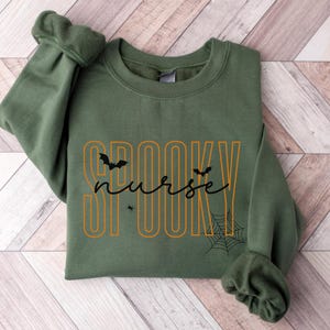 Puede incluir: Una sudadera verde oliva con la frase "Spooky Nurse" en letras naranjas y negras. El dise&ntilde;o incluye murci&eacute;lagos, una ara&ntilde;a y una telara&ntilde;a, ideal para Halloween. La sudadera est&aacute; colocada sobre una superficie de madera.