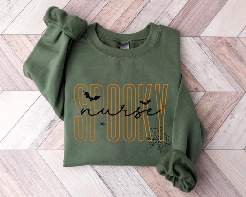 Puede incluir: Una sudadera verde oliva con la frase "Spooky Nurse" en letras naranjas y negras. El dise&ntilde;o incluye murci&eacute;lagos, una ara&ntilde;a y una telara&ntilde;a, ideal para Halloween. La sudadera est&aacute; colocada sobre una superficie de madera.
