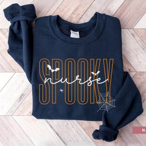 Puede incluir: Sudadera azul marino con las palabras "SPOOKY nurse" en naranja y blanco. El dise&ntilde;o incluye murci&eacute;lagos y una telara&ntilde;a, perfecto para Halloween. La sudadera es de cuello redondo.