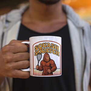 Könnte beinhalten: Weiße Keramik-Tasse mit dem Text "Squash Squatch" in Orange und Gelb, mit einem Cartoon-Sasquatch, der einen Squash-Schläger hält. Das Design enthält Retro-Streifen. Ein lustiger Artikel.