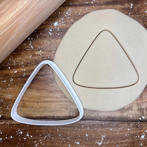 May include: A white plastic triangle cookie cutter next to a triangle cut-out in dough. A wooden rolling pin is in the upper left corner. Flour dusts the wooden surface.