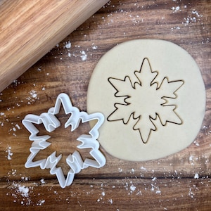 May include: A white plastic snowflake cookie cutter next to a cut-out snowflake in dough. A wooden rolling pin is in the background, with flour sprinkled on the wooden surface. The cookie cutter is approximately 4 inches in diameter.