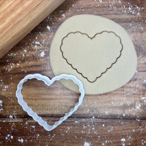 May include: A white, scalloped-edge heart-shaped cookie cutter next to a cut-out heart in a piece of dough. A wooden rolling pin and flour are also visible on the wooden surface. The image suggests baking or crafting.