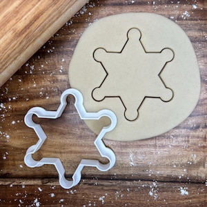 Pode incluir: Um cortador de biscoitos branco em forma de estrela de xerife ao lado de um recorte da mesma forma em um círculo de massa. Um rolo de madeira está no fundo sobre uma superfície de madeira.