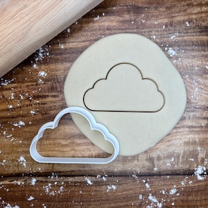 Può includere: Un tagliabiscotti bianco a forma di nuvola accanto a un ritaglio a forma di nuvola in un pezzo rotondo di pasta. Un mattarello di legno è sullo sfondo. La scena è ambientata su una superficie di legno cosparsa di farina.