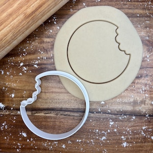 Puede incluir: Un cortador de galletas de plástico blanco en forma de círculo con un mordisco, junto a un círculo de masa para galletas con un mordisco. Un rodillo de madera está en el fondo.
