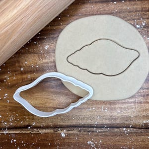 Puede incluir: Un cortador de galletas de plástico blanco con forma de concha junto a una forma de concha recortada en un círculo de masa. Un rodillo de madera está en el fondo sobre una superficie de madera.