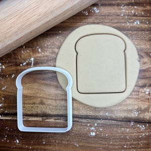 May include: A white plastic bread-shaped cookie cutter beside a piece of dough with a bread imprint. A wooden rolling pin is in the background, and flour is sprinkled on the wooden surface. The items are for baking.