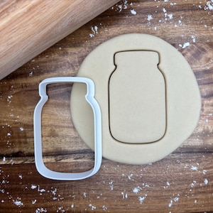 Può includere: Un tagliabiscotti bianco a forma di barattolo di vetro accanto a un biscotto ritagliato su una superficie di legno. Un mattarello è visibile sullo sfondo. Il tagliabiscotti è vuoto e il biscotto è marrone chiaro.