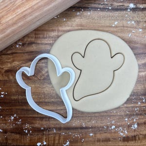 May include: A white plastic ghost-shaped cookie cutter next to a cut-out ghost shape in a round piece of dough. A wooden rolling pin is in the background on a wooden surface. Flour is sprinkled around the dough.