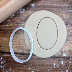 May include: A white oval cookie cutter next to a cut-out oval shape in a piece of dough. A wooden rolling pin is in the background, with flour sprinkled on a wooden surface. Baking and cookie-making concept.