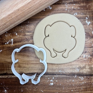 May include: A white plastic turkey-shaped cookie cutter next to a cut-out cookie and a wooden rolling pin. The cookie cutter and cookie are on a wooden surface, with flour sprinkled around.