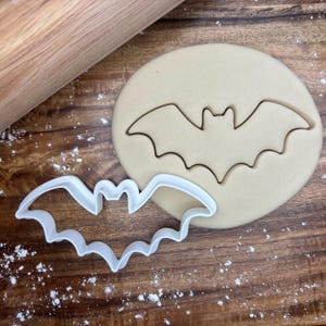 Puede incluir: Un cortador de galletas de plástico blanco con forma de murciélago junto a un trozo redondo de masa con una huella de murciélago. Un rodillo de madera y una superficie de madera están en el fondo. Harina espolvoreada.