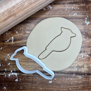 May include: A white plastic bird-shaped cookie cutter next to a cutout of a bird in dough. A wooden rolling pin is in the background. The dough is on a wooden surface with flour.