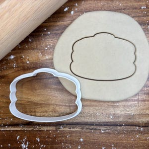 May include: A white plastic cookie cutter in the shape of a stack of pancakes sits next to a circle of dough with the pancake outline pressed into it. A wooden rolling pin is in the upper left corner.