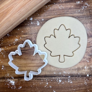 May include: A white plastic maple leaf cookie cutter next to a cut-out maple leaf in a circle of dough. A wooden rolling pin is in the background. Flour is sprinkled on the wooden surface.