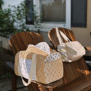 May include: A tan and cream-colored duffel bag with a floral pattern and cream-colored handles sits on a wooden chair. A tote bag with a floral pattern and a white strap is also on the chair. A white and black polka dot bag is inside the duffel bag.