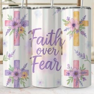 May include: A cylindrical tumbler featuring a watercolour design of pastel crosses adorned with purple, pink, and yellow flowers. The text Faith over Fear is printed in a purple script font. The background has faint vertical stripes and floral accents.