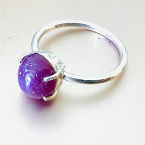 May include: A silver ring featuring a large, oval-shaped, deep purple gemstone. The gemstone is held in place by four silver prongs. The ring band is a simple, polished silver. The ring is set against a light background.
